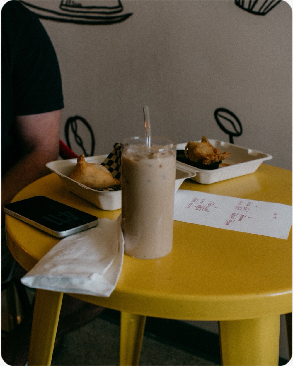 A glass of iced chai, a samosa and pakoras on a yellow table beside a handwritten menu